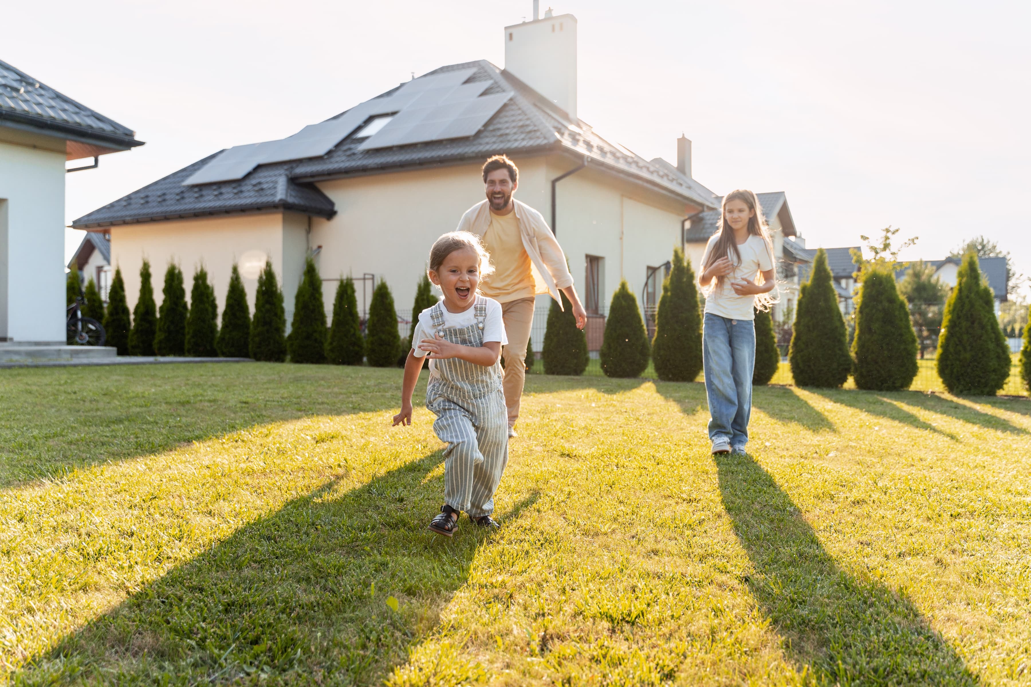Glückliche Familie spielt im Garten vor Haus mit Photovoltaikanlage in der Abendsonne